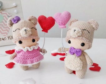 May include: Two handmade crochet teddy bears, one in a pink dress and the other with a purple bow tie, holding heart-shaped balloons. The bears are beige with black eyes and are surrounded by red heart-shaped confetti.
