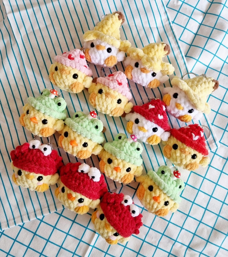 May include: A collection of 13 crocheted chick keychains with various hats. The hats include a mushroom, a witch's hat, a frog, and a red hat with googly eyes. The chicks are all yellow and have black eyes.