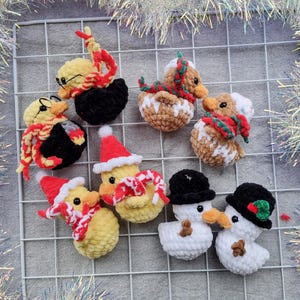 May include: A collection of handmade crochet Christmas ornaments. The ornaments include yellow ducks with Santa hats and scarves, snowmen with black hats, and ducks with glasses and scarves. The ornaments are arranged on a white grid.