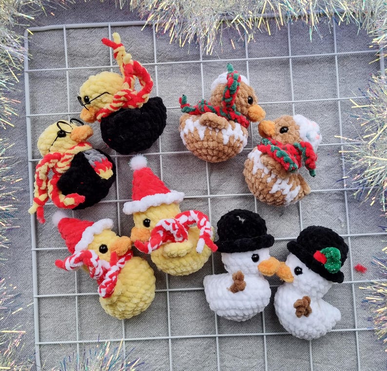 May include: A collection of handmade crochet Christmas ornaments. The ornaments include yellow ducks with Santa hats and scarves, snowmen with black hats, and ducks with glasses and scarves. The ornaments are arranged on a white grid.