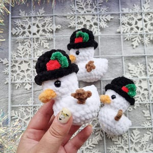 May include: Crocheted snowman ornaments. Each snowman is white with a black hat adorned with red and green accents, an orange beak, and a brown twig-like arms. The ornaments are handmade and suitable for holiday decor.