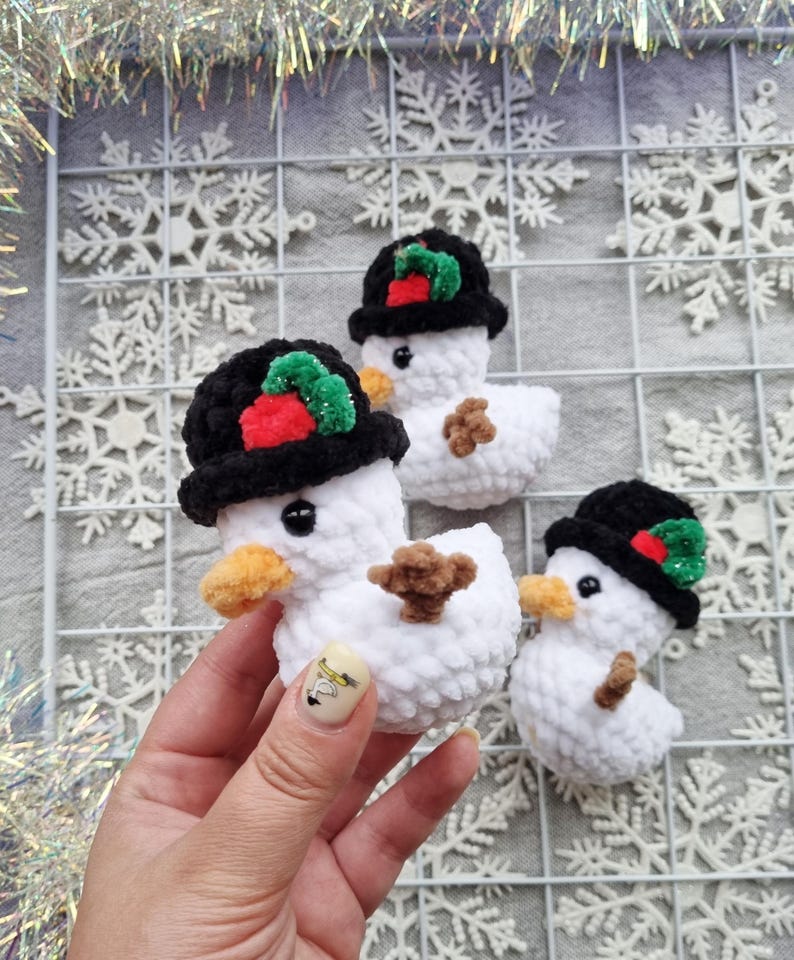 May include: Crocheted snowman ornaments. Each snowman is white with a black hat adorned with red and green accents, an orange beak, and a brown twig-like arms. The ornaments are handmade and suitable for holiday decor.