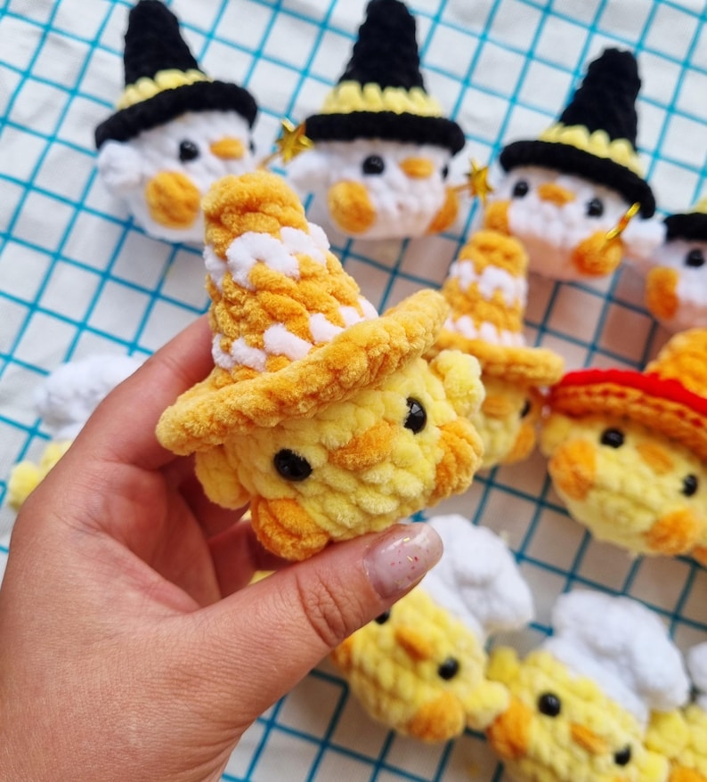 May include: A collection of small, crocheted plush figures made from soft yellow and white yarn. Each figure features black bead eyes and wears a different colored hat, including black witch hats, yellow and white striped hats, and red-brimmed hats.