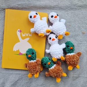 May include: A collection of handmade crochet ducklings and a yellow book with a goose illustration. The ducklings are white and brown with orange beaks and yellow feet. The book has the word "Wow" on it.