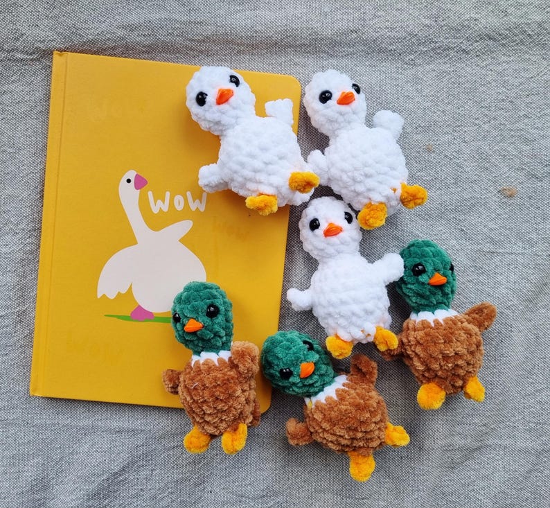 May include: A collection of handmade crochet ducklings and a yellow book with a goose illustration. The ducklings are white and brown with orange beaks and yellow feet. The book has the word "Wow" on it.