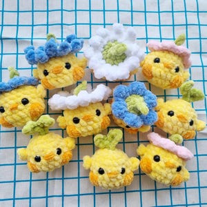 May include: A collection of handmade crochet chick ornaments in various colors. The chicks are primarily yellow with orange accents and black eyes. Some chicks have blue, white, or pink floral headwear, and green leaf accents. The ornaments are arranged on a blue and white checkered fabric.