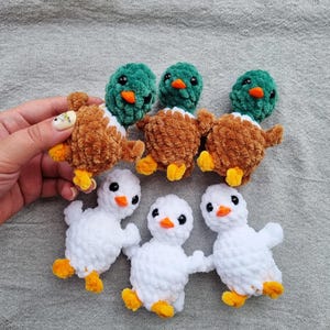 May include: A collection of handmade crochet ducklings and ducklings. The ducks have brown and white bodies with green heads and orange beaks. The ducklings are white with orange feet and beaks. The toys are arranged on a gray fabric surface.
