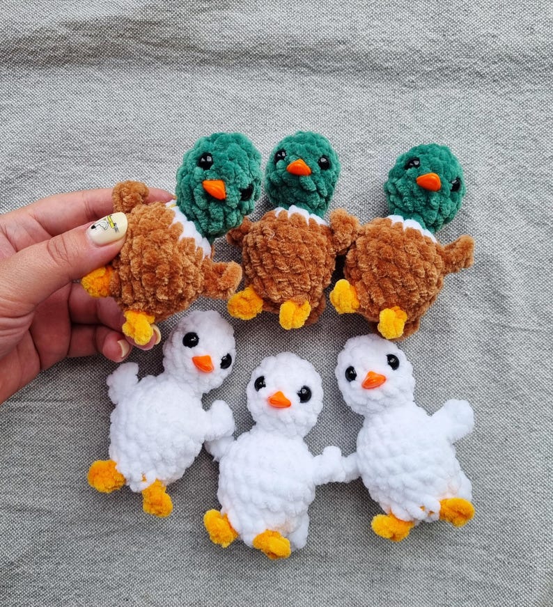 May include: A collection of handmade crochet ducklings and ducklings. The ducks have brown and white bodies with green heads and orange beaks. The ducklings are white with orange feet and beaks. The toys are arranged on a gray fabric surface.