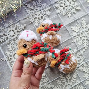 May include: Three handmade crochet duck ornaments. The ducks are brown, white, and tan with red and green scarves. Each duck has a white hat with pink sprinkles. The ducks are on a white grid with snowflake decorations.