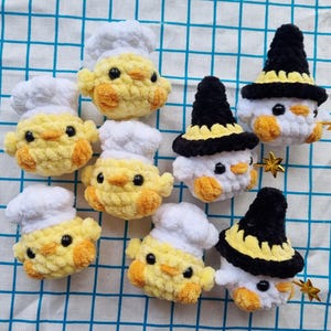 May include: A collection of seven small, crocheted plush figures. Four yellow chicks wear white chef hats, while three white figures wear black and yellow pointed hats. Each figure has black bead eyes and small orange beaks and feet.