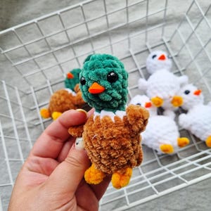 May include: A handmade, plush duckling with a green head, brown body, and orange beak and feet. The duckling has black safety eyes and a white collar. Other similar ducklings are in the background, some white and some brown.
