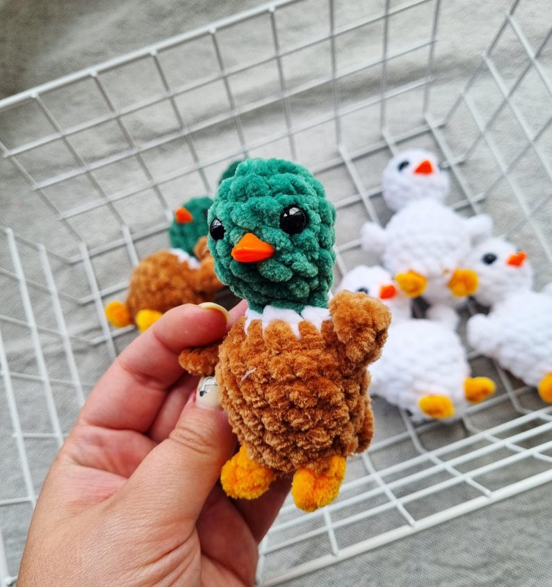 May include: A handmade, plush duckling with a green head, brown body, and orange beak and feet. The duckling has black safety eyes and a white collar. Other similar ducklings are in the background, some white and some brown.