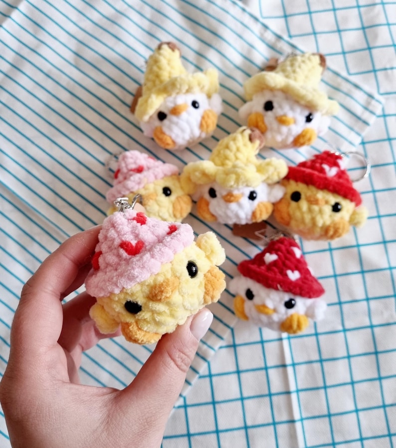 May include: A collection of six crocheted chick keychains. Each keychain features a yellow chick with a different colored hat. The hats include yellow, pink, red, and white. The keychains are perfect for adding a touch of whimsy to your keys or bag.