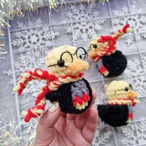 May include: Crocheted duck figurines, each with a yellow head, orange beak, and black glasses. The ducks wear red and yellow scarves and black robes. One is held in a hand, and two others are on a white grid background.