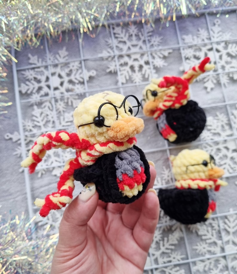 May include: Crocheted duck figurines, each with a yellow head, orange beak, and black glasses. The ducks wear red and yellow scarves and black robes. One is held in a hand, and two others are on a white grid background.