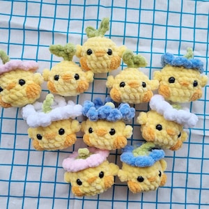 May include: A collection of handmade yellow crochet chick ornaments with various colored accessories, including pink, blue, and green accents. The chicks have black eyes and orange cheeks, and are arranged on a blue and white checkered surface.