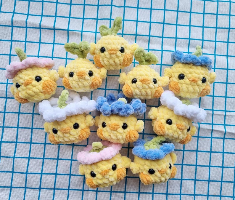May include: A collection of handmade yellow crochet chick ornaments with various colored accessories, including pink, blue, and green accents. The chicks have black eyes and orange cheeks, and are arranged on a blue and white checkered surface.
