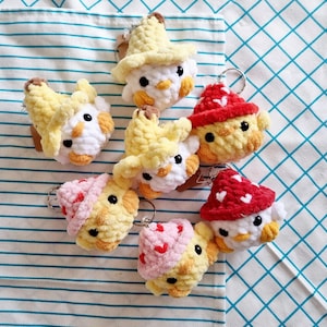 May include: A set of seven crocheted chick keychains. Each keychain features a different colored hat, including red, yellow, and pink. The chicks have white faces and yellow bodies.