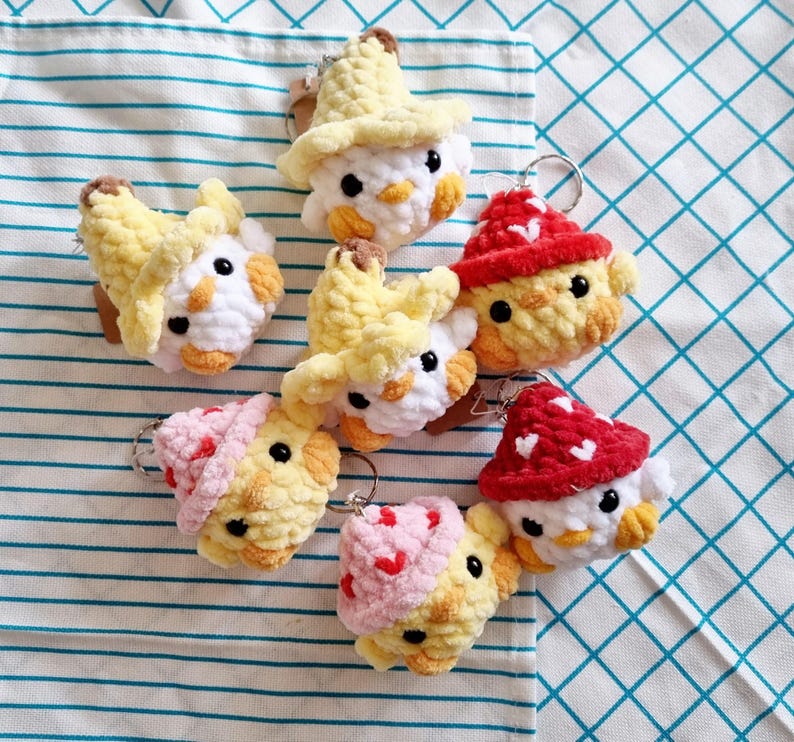 May include: A set of seven crocheted chick keychains. Each keychain features a different colored hat, including red, yellow, and pink. The chicks have white faces and yellow bodies.