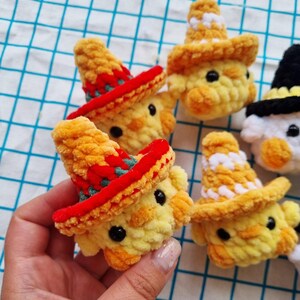 May include: Several small, round, yellow crocheted chick figures with black bead eyes and orange beaks. Some chicks wear crocheted hats in red with green accents, while others wear yellow and white hats. One chick wears a black hat.