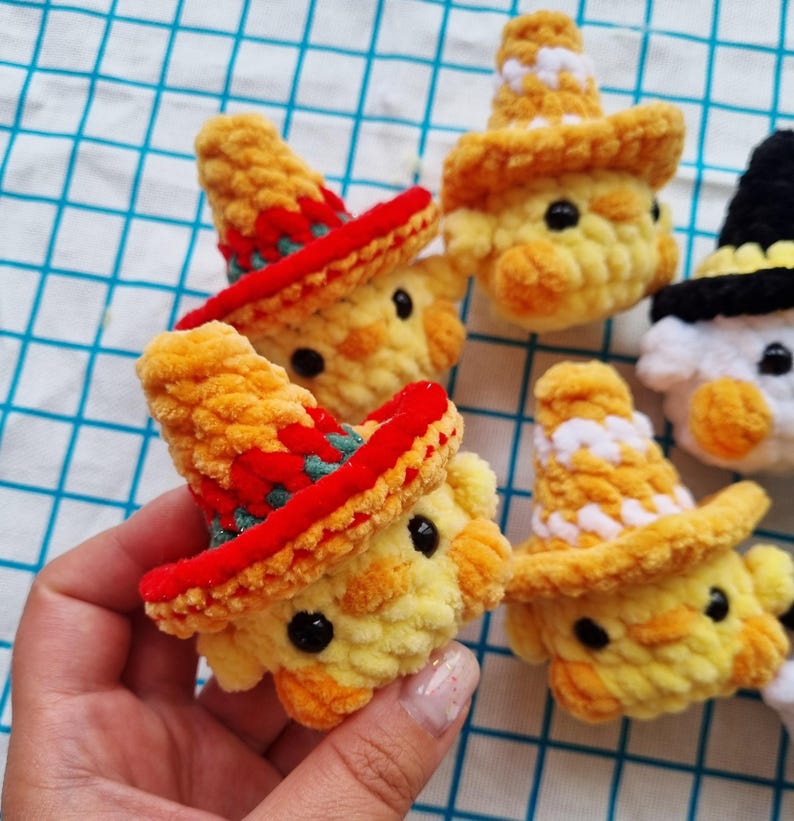 May include: Several small, round, yellow crocheted chick figures with black bead eyes and orange beaks. Some chicks wear crocheted hats in red with green accents, while others wear yellow and white hats. One chick wears a black hat.