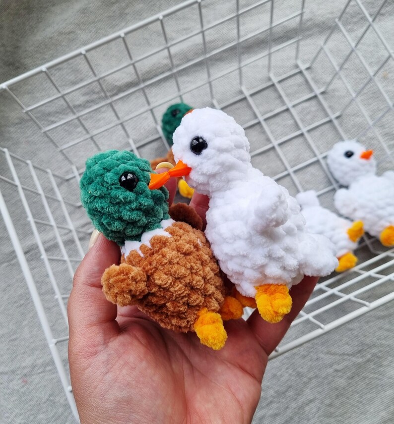 May include: Crocheted ducklings in various colors. One duckling is white with orange beak and yellow feet, another is green and brown. The ducklings are handmade and fluffy, with black eyes. The background includes a white wire basket.