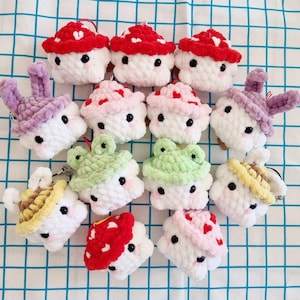 Puede incluir: Una colección de 12 juguetes de peluche tejidos a crochet en varios colores y formas, incluyendo hongos, ranas y conejos. Cada juguete tiene una cara blanca con ojos negros y una pequeña boca.