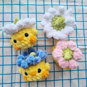 May include: Four handmade crochet items: two yellow chick-like figures with cloud and flower accents, and two flower-shaped items in white and pink. The items are made of soft, textured yarn and sit on a blue and white checkered surface.