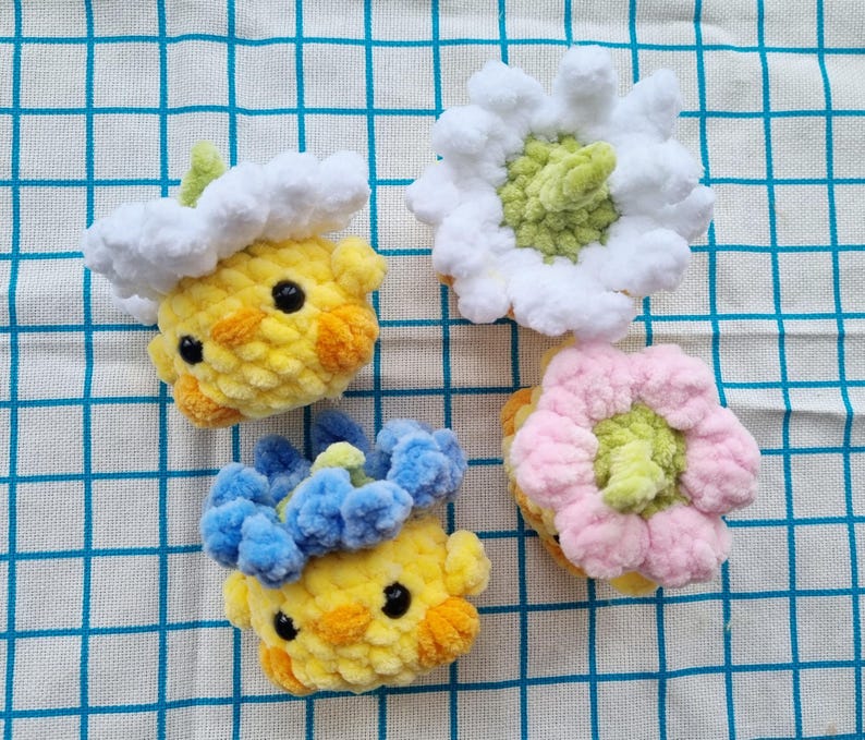 May include: Four handmade crochet items: two yellow chick-like figures with cloud and flower accents, and two flower-shaped items in white and pink. The items are made of soft, textured yarn and sit on a blue and white checkered surface.