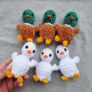 May include: Six handmade crochet ducklings. Three ducks have green heads, brown bodies, and yellow feet. The other three are white with orange beaks and yellow feet. The ducklings are arranged on a gray surface.