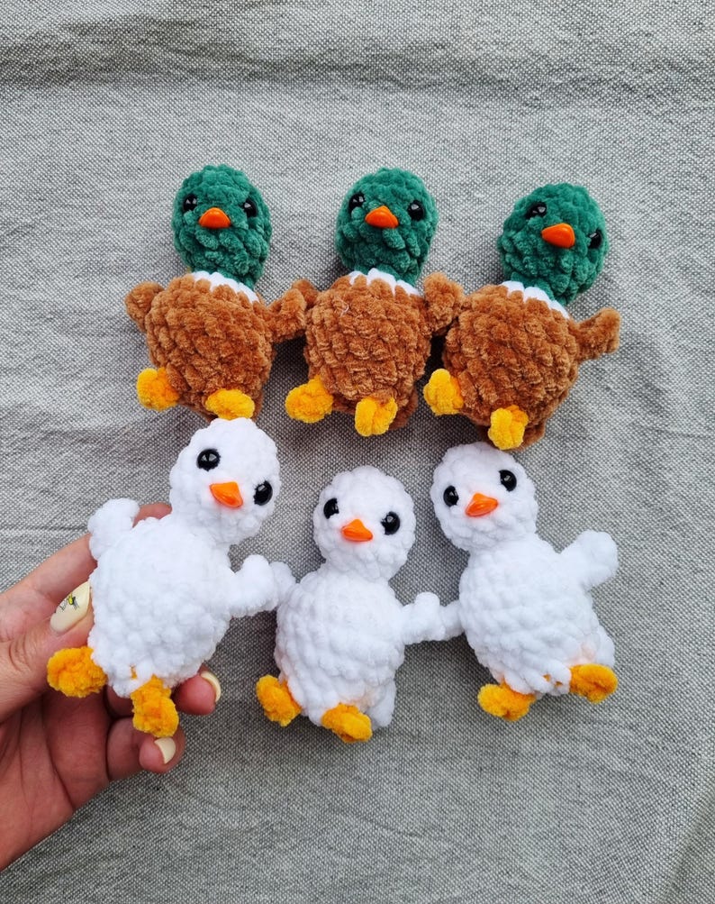 May include: Six handmade crochet ducklings. Three ducks have green heads, brown bodies, and yellow feet. The other three are white with orange beaks and yellow feet. The ducklings are arranged on a gray surface.