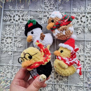 May include: Four handmade crochet duck ornaments. One is a snowman, another is wearing a gingerbread hat and scarf, a third is dressed as a wizard, and the last is wearing a Santa hat and scarf. The ducks are on a white snowflake background.