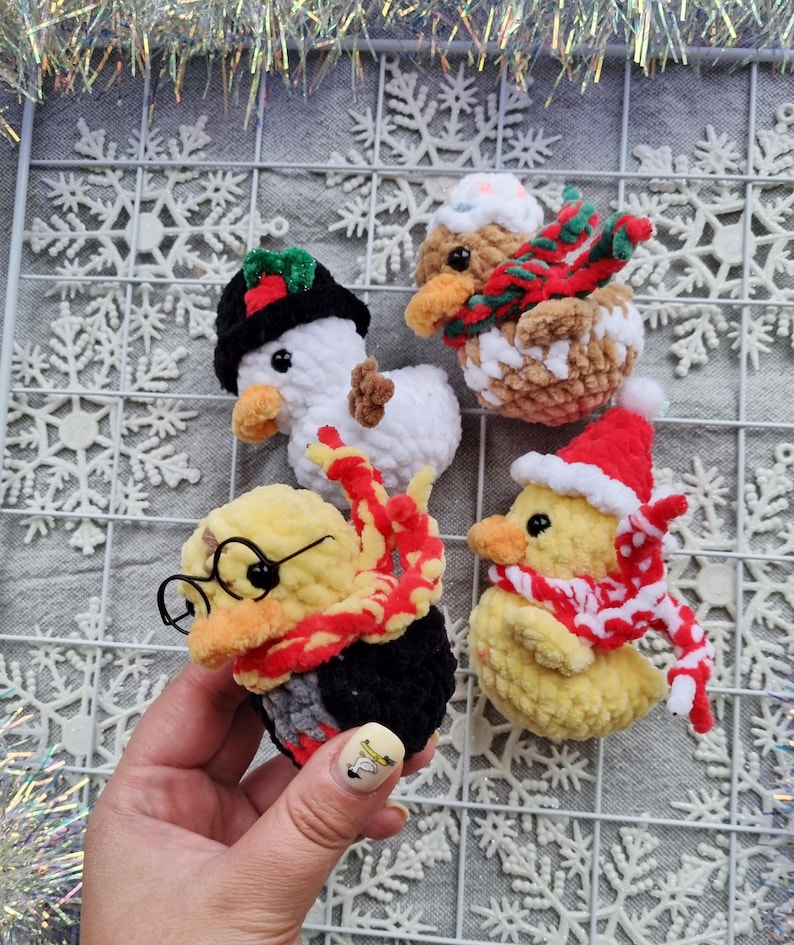 May include: Four handmade crochet duck ornaments. One is a snowman, another is wearing a gingerbread hat and scarf, a third is dressed as a wizard, and the last is wearing a Santa hat and scarf. The ducks are on a white snowflake background.
