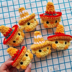 May include: Five small, yellow, crocheted plush chicks, each wearing a miniature sombrero. The sombreros are primarily orange with red and green accents, or white and orange patterns. Each chick features two small, round, black plastic eyes.