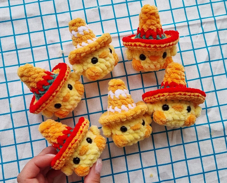 May include: Five small, yellow, crocheted plush chicks, each wearing a miniature sombrero. The sombreros are primarily orange with red and green accents, or white and orange patterns. Each chick features two small, round, black plastic eyes.