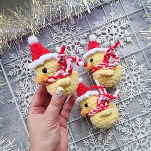 May include: Three handmade yellow crochet ducklings wearing red Santa hats and red and white striped scarves. The ducklings have black eyes and orange beaks. The background includes snowflake decorations and silver tinsel.