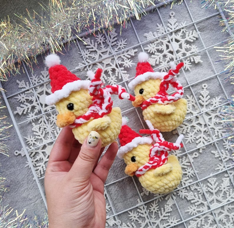 May include: Three handmade yellow crochet ducklings wearing red Santa hats and red and white striped scarves. The ducklings have black eyes and orange beaks. The background includes snowflake decorations and silver tinsel.