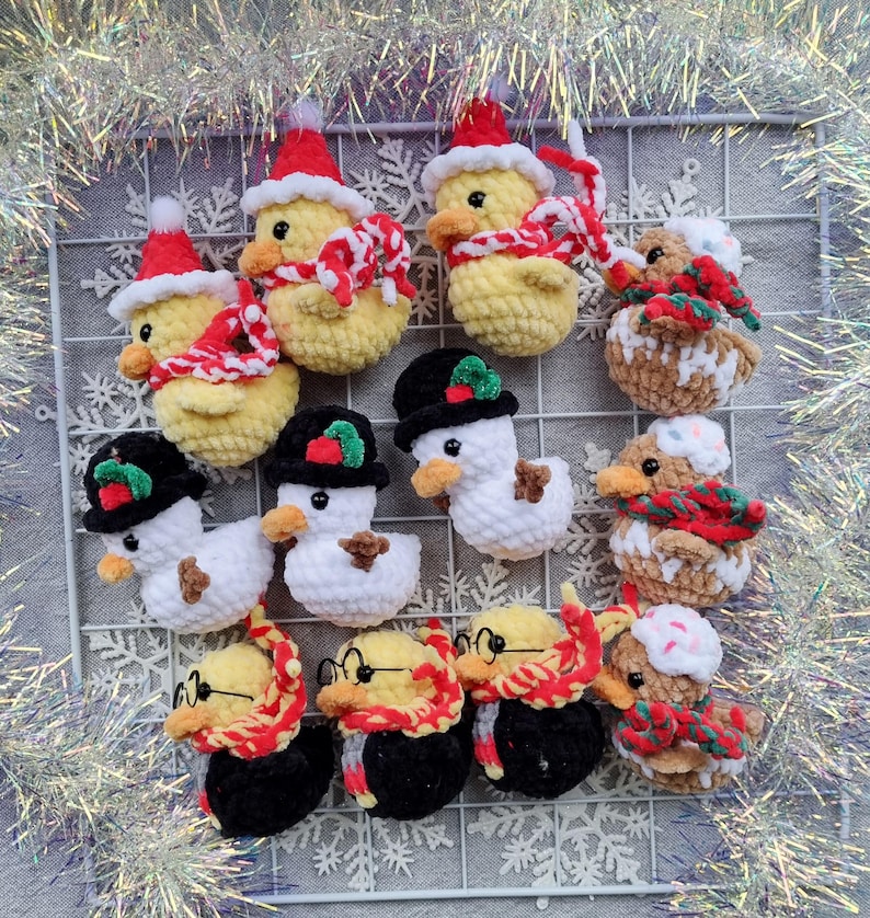 May include: A collection of handmade crochet duck ornaments in various festive designs. Some ducks are yellow with red Santa hats and scarves, while others are white snowmen with black hats. Other ducks wear glasses and scarves.