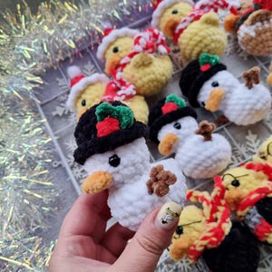 May include: A collection of handmade crochet Christmas ornaments. The ornaments include snowmen with black hats and ducks wearing Santa hats and scarves. The snowmen have brown twig arms and orange beaks. The ducks are yellow with red and white scarves.