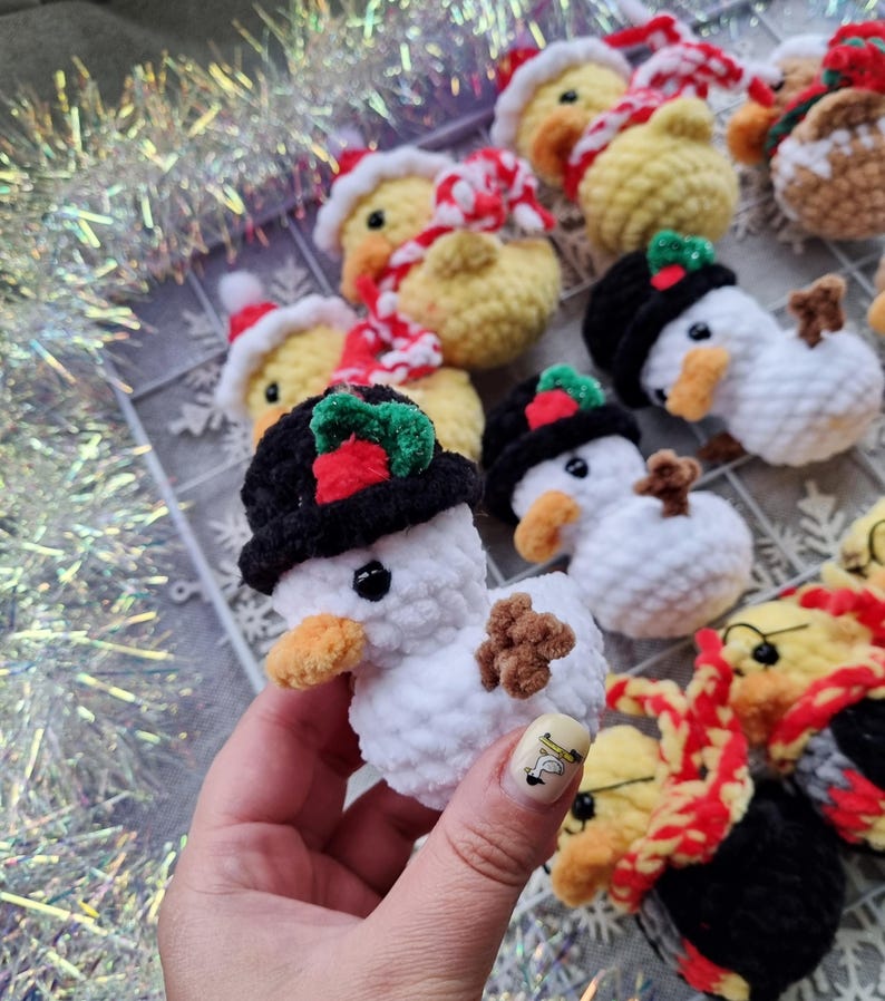 May include: A collection of handmade crochet Christmas ornaments. The ornaments include snowmen with black hats and ducks wearing Santa hats and scarves. The snowmen have brown twig arms and orange beaks. The ducks are yellow with red and white scarves.