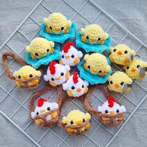 May include: A selection of handmade crochet chick and chicken-themed items. The chicks are yellow and on blue pads, while the chickens have red combs. Some are keyrings, others are in brown baskets, all on a white grid.