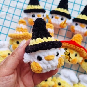 May include: A collection of small, crocheted plush figures shaped like ducks, each wearing a different hat. One prominent white duck features a black and yellow pointed hat and holds a small, gold-colored star-shaped wand in its wing.