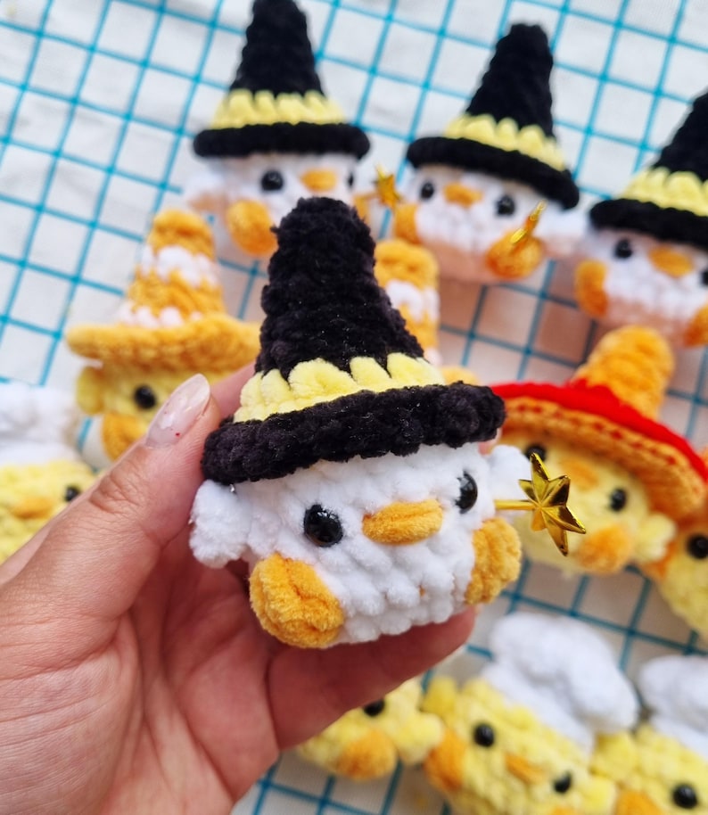 May include: A collection of small, crocheted plush figures shaped like ducks, each wearing a different hat. One prominent white duck features a black and yellow pointed hat and holds a small, gold-colored star-shaped wand in its wing.