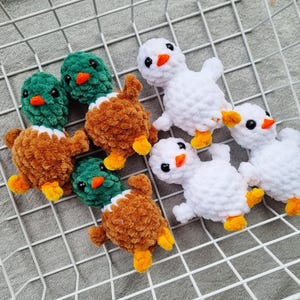 May include: Handmade crochet ducklings in white, brown, and green. The ducklings have orange beaks and feet, and black eyes. The toys are arranged on a white wire grid, perfect for a nursery or as a gift.