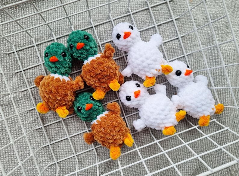 May include: Handmade crochet ducklings in white, brown, and green. The ducklings have orange beaks and feet, and black eyes. The toys are arranged on a white wire grid, perfect for a nursery or as a gift.