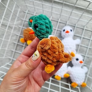 May include: Close-up of several handmade crochet ducklings. One duckling is in focus, featuring a brown body, green head, orange beak, and yellow feet. Other ducklings are visible, some brown and others white, all with similar features.