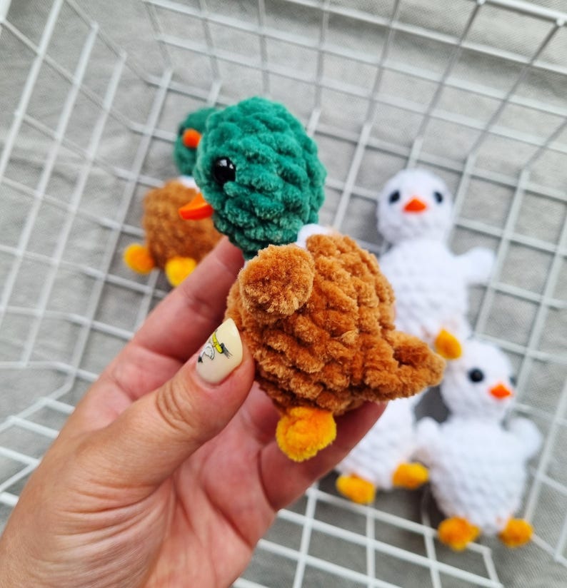 May include: Close-up of several handmade crochet ducklings. One duckling is in focus, featuring a brown body, green head, orange beak, and yellow feet. Other ducklings are visible, some brown and others white, all with similar features.