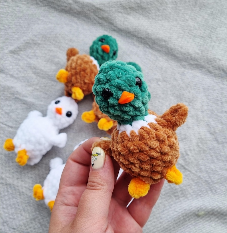 May include: A collection of handmade crochet ducklings in various colors. The ducklings have brown, green, and white bodies with orange beaks and yellow feet. The ducklings are small and fluffy, perfect for a child's toy or nursery decor.