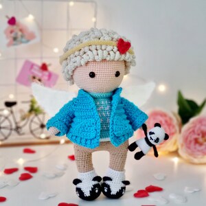 Peut inclure: Une poupée ange en crochet portant un pull bleu, des ailes blanches et un cœur rouge sur la tête. La poupée tient un petit panda en crochet.