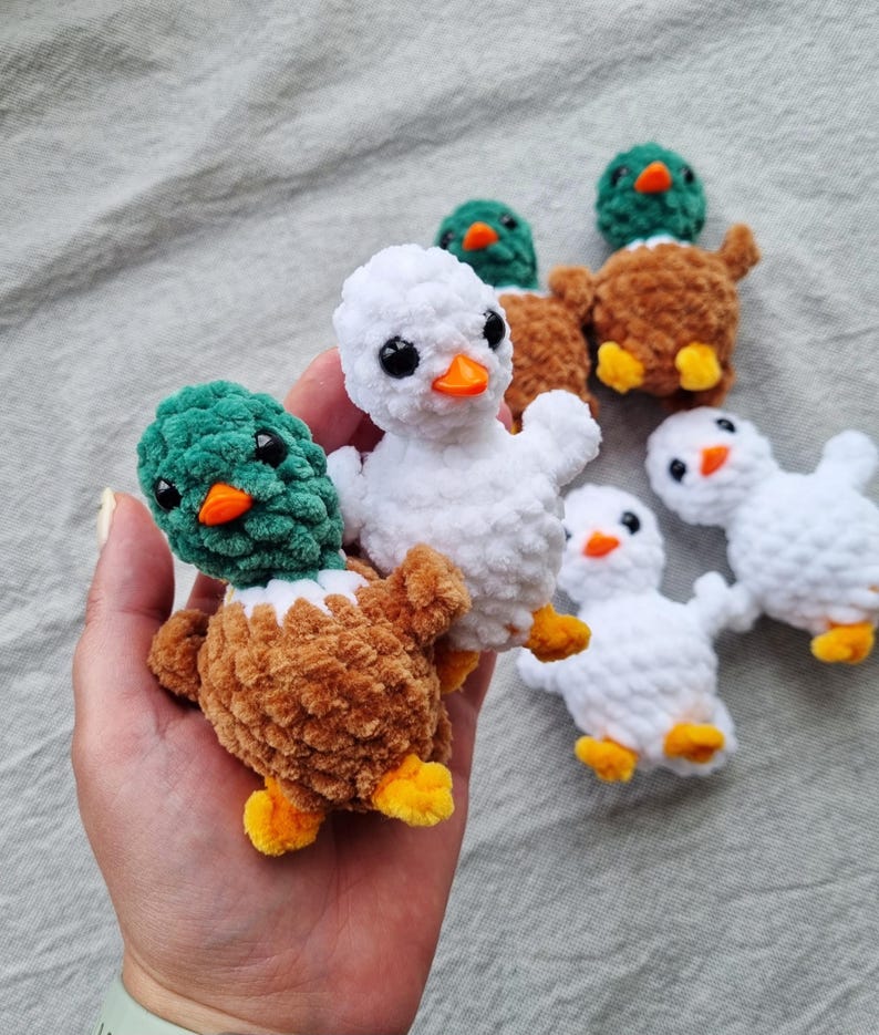 May include: A collection of handmade crochet ducklings in various colors. The ducklings feature white, brown, and green bodies with orange beaks and yellow feet. The plush toys are soft and cuddly, perfect for children.